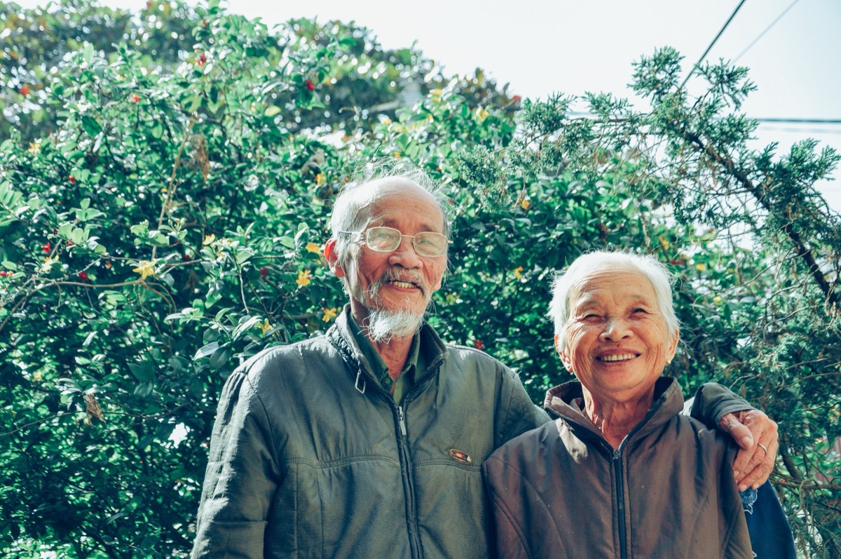 An older couple standing together outdoors in front of greenery.