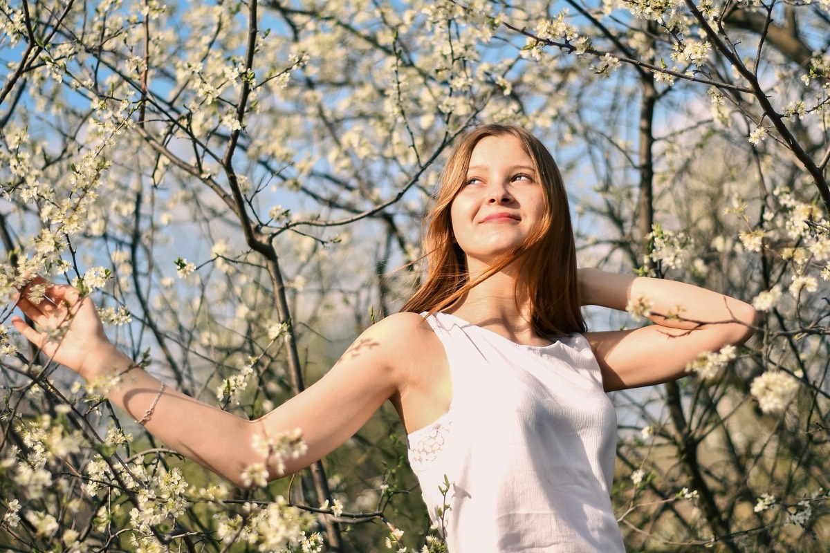 A woman standing outdoors with her arms open beneath blossoming branches.
